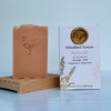 Bar of soap with rooster design on a wooden stand next to its packaging labeled 'Woodland Sunrise'.
