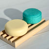 Two bars of soap, one yellow and one turquoise, on a wooden soap dish against a light background.