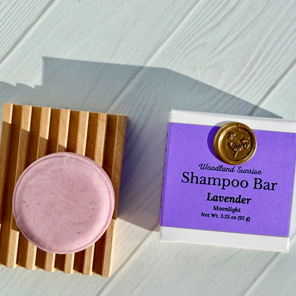 Lavender shampoo bar on a wooden soap dish with its packaging on a light wooden surface.