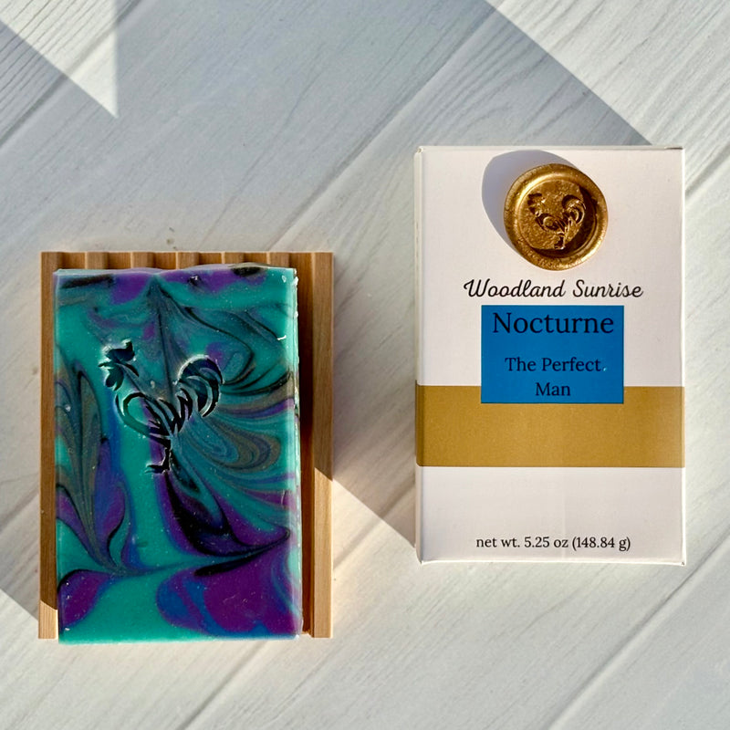 Bar of soap with a decorative design on a wooden stand next to its packaging labeled 'Woodland Sunrise Nocturne'.