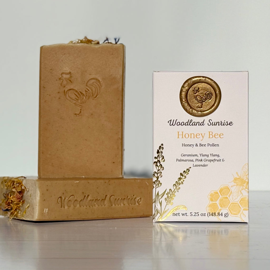 Bar of soap and packaging labeled 'Woodland Sunrise Honey Bee' on a reflective surface with a light gray background.