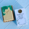 Bar of soap with green and gold design next to a box labeled 'Woodland Sunrise Spa Day' on a blue wooden surface.