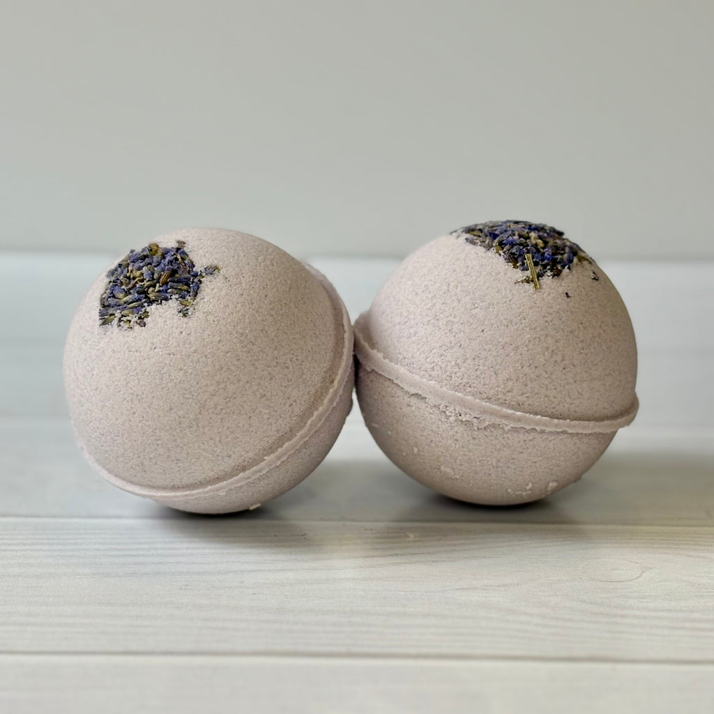 Two pale purple bath bombs with lavender on a light wooden surface