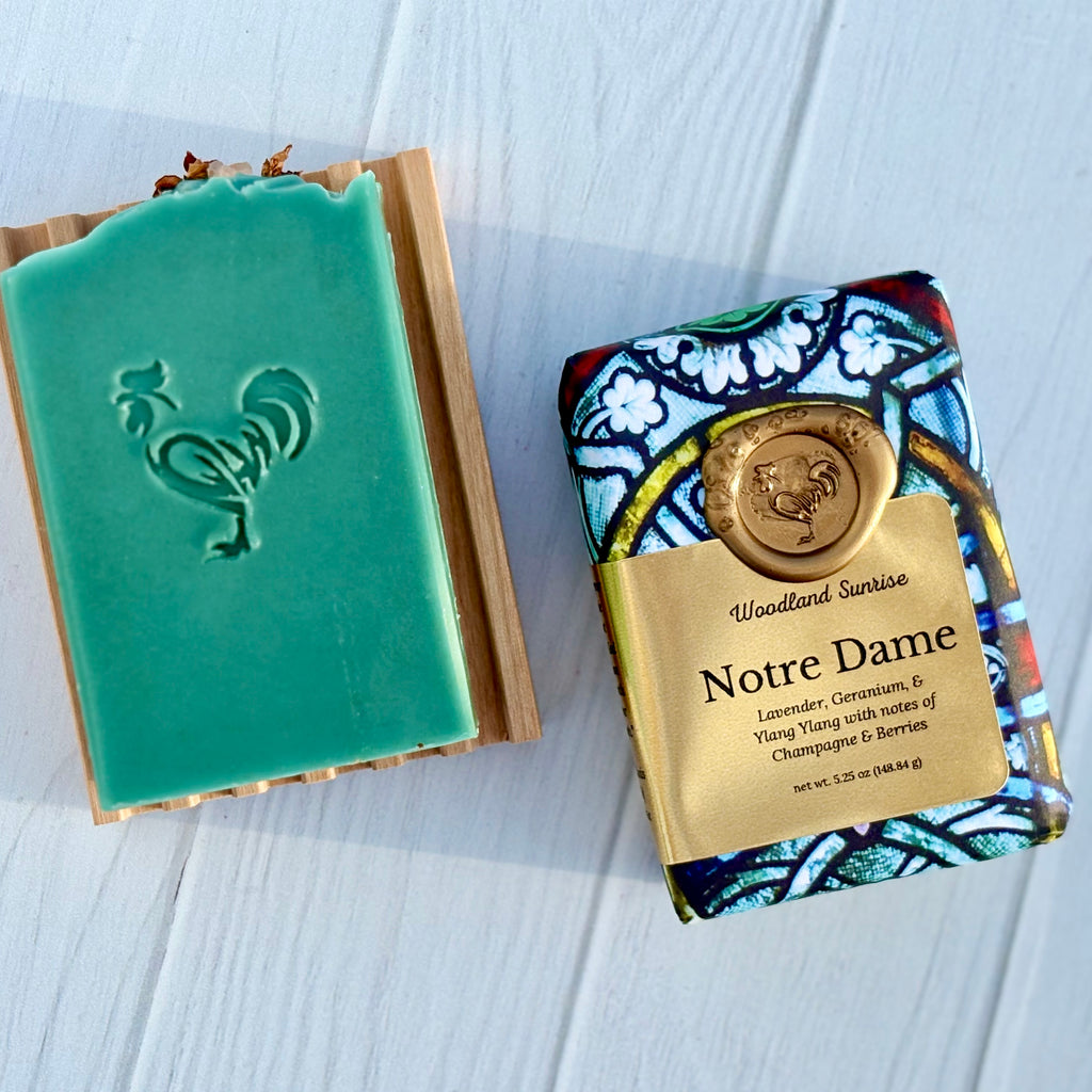 Green soap bar with rooster stamp and colorful packaging labeled 'Notre Dame' on a white wooden surface.