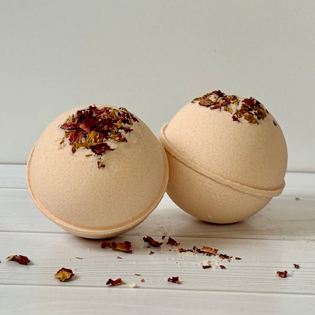 Two pale pink bath bombs with dried herbs on a light wooden surface