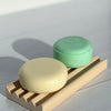 Two round soap bars, one beige and one green, on a wooden soap dish against a light background.