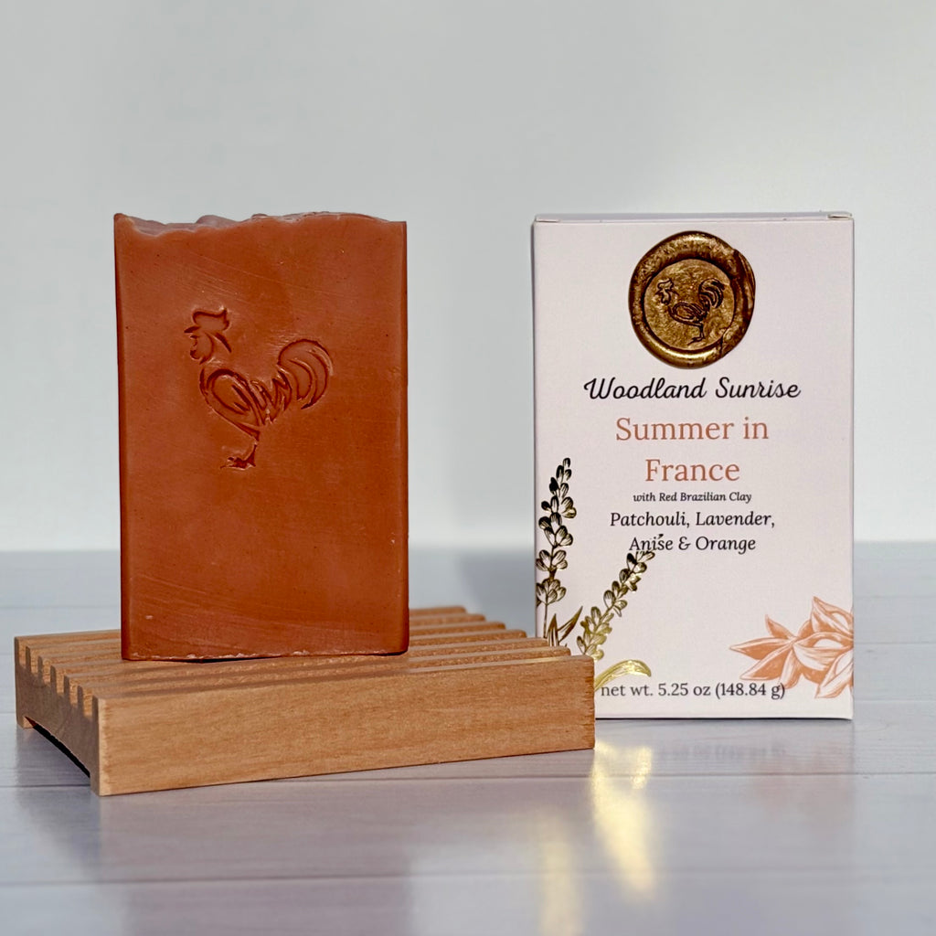 Brown soap bar with rooster design on a wooden stand next to its packaging labeled 'Woodland Sunrise Summer in France'.